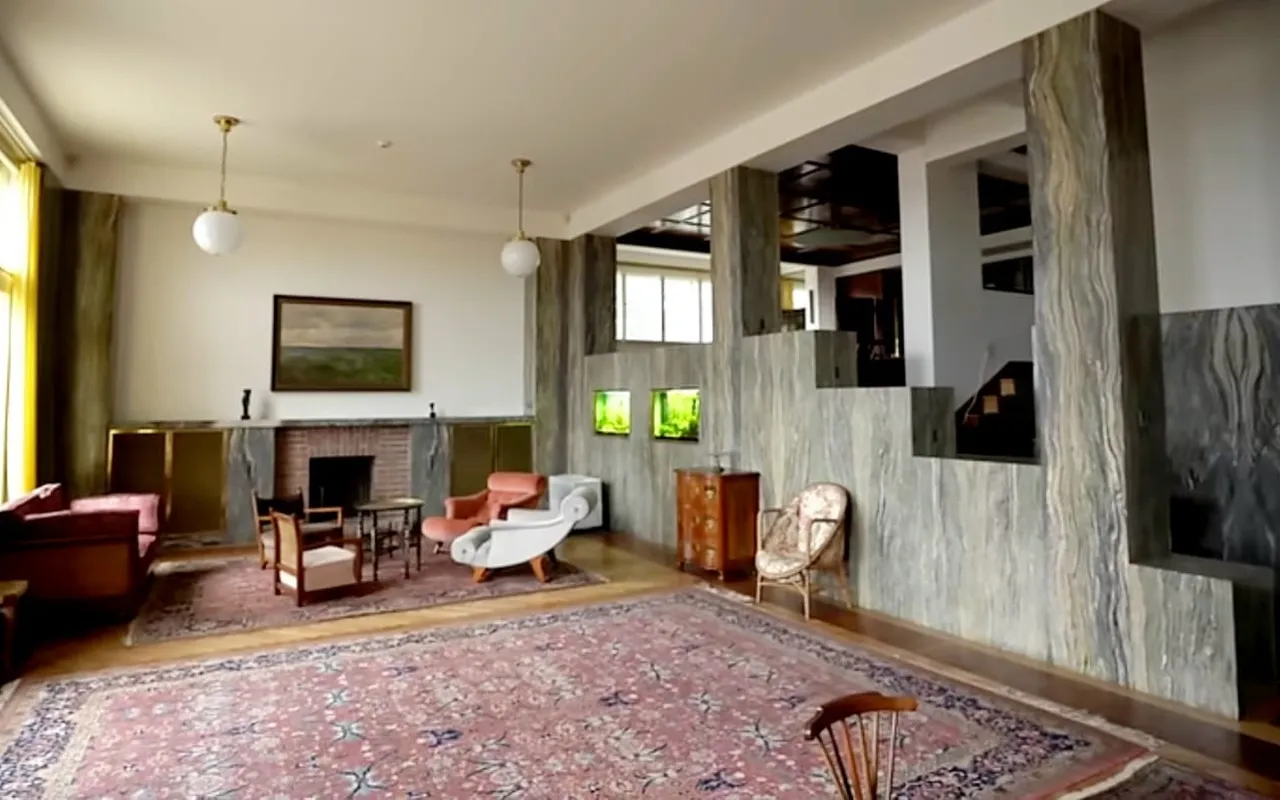Il salone di Villa Müller con la sua parete in marmo Cipollino verde, il camino e i tappeti, la sala da pranzo che si apre al piano superiore